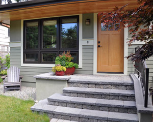The porch is built with concrete footings, CMU, and finished with pavers and caps, and stucco on the vertical sides.  Stucco in a darker green color is also part of the siding, which includes Hardiplank siding at the lower section, divided by a wood belly band. The new fiberglass entry door and natural wood soffits completes the look